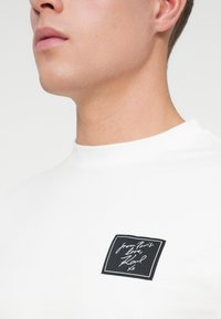 Suur lähivõte inimesest, kes kannab valget T-särki, mille rinnal on must ruudukujuline plaastriga, millel on kiri "Pariisi armastusest, Karl xx".