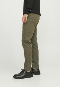 Chinos verde oliva con un corte ajustado, hechos de una mezcla de algodón; cuenta con bolsillos laterales y un bolsillo trasero discreto; llevados con zapatos negros.