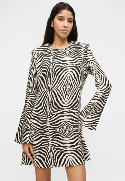 Γυναίκα με μακριά σκούρα μαλλιά φορώντας μαύρο και λευκό μίνι φόρεμα με γεωμετρικό μοτίβο, μακριά μανίκια και δομημένους ώμους, στέκεται μπροστά σε ένα απλό φόντο.