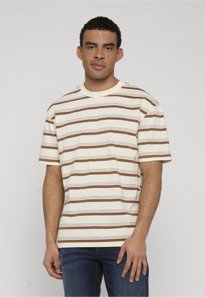 Młody mężczyzna z krótkimi kręconymi włosami, ubrany w beżowo-brązową koszulę w paski z krótkim rękawem oraz niebieskie dżinsy, stoi na tle jednolitego tła.