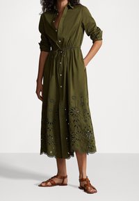 Olivengrön, långärmad skjortklänning med knappar, insnörd midja och blommigt utskuret detalj vid fållen, parat med bruna remsandaler.