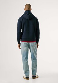 Sudadera con capucha color navy, con puños y dobladillo acanalados, superpuesta a una camiseta roja, combinada con vaqueros de un azul claro y zapatos informales.