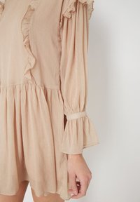 Vestido beige claro con una tela texturizada, con bordes arrugados en el escote y las mangas, y detalles de fruncido en la cintura.