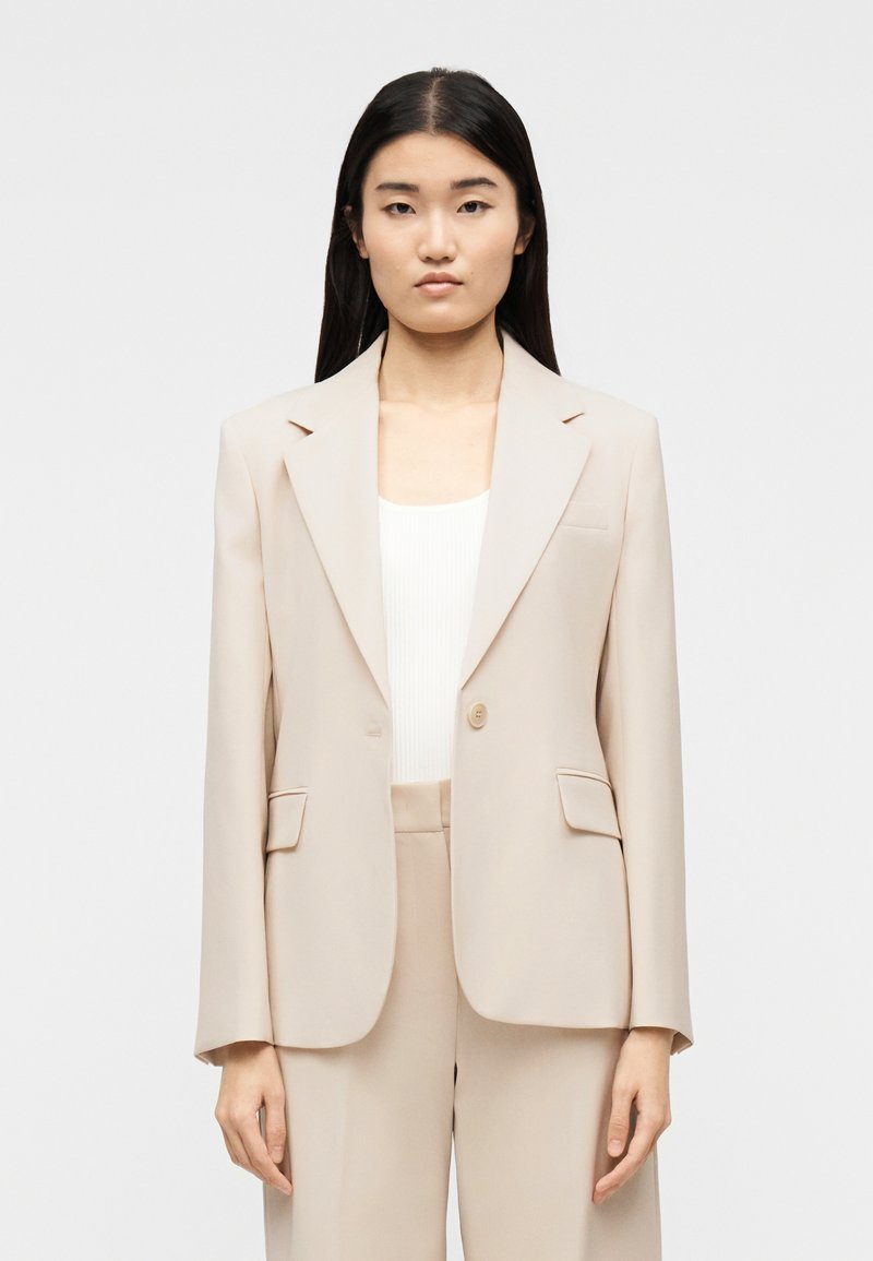 Γυναίκα με μακριά σκουρόχρωμα μαλλιά που φοράει μπεζ σακάκι και παντελόνι που ταιριάζουν, πάνω από λευκή ριμπ πλεκτή μπλούζα, στέκεται μπροστά σε απλό λευκό φόντο.