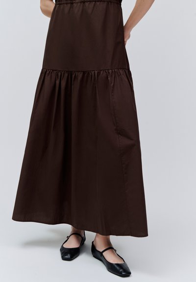 Longue jupe marron foncé avec une couture froncée au-dessus du mollet, portée avec des ballerines Mary Jane noires sur un fond clair.