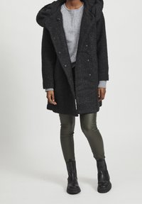 Svart texturerad jacka med huva, featuring stora knappar och en mjuk krage, matchad med mörkgröna åtsittande byxor och svarta ankelstövlar.