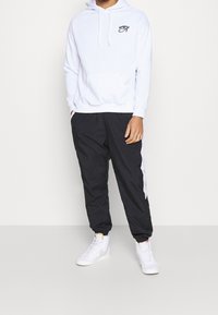 Uomo che indossa una felpa con cappuccio bianca con il logo nero dell'Occhio di Horus, pantaloni sportivi neri con striscia bianca e scarpe da ginnastica bianche alte, in piedi contro un muro bianco.