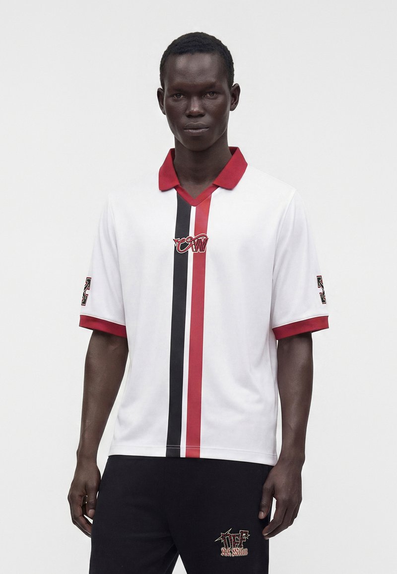 Homme portant une chemise blanche à manches courtes avec des rayures verticales rouges et noires et un col rouge, associée à un pantalon noir avec un texte graphique sur la cuisse.