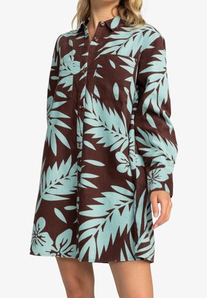 Frau trägt ein langärmliges braunes Kleid mit großen hellblauen tropischen Blatt- und Blumenmustern, Knöpfen vorne und Brusttaschen.