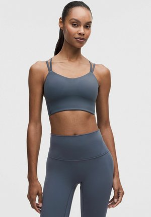 Mujer de pie con sujetador deportivo gris oscuro a juego con doble tirante y leggings de talle alto sobre fondo liso.