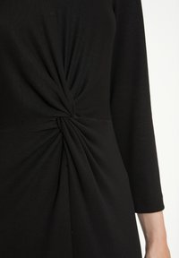 Haut noir avec des manches trois-quarts, un détail à nœud tordu à la taille et une texture lisse et extensible.