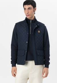 Marineblå quilted vest med trykknapper, to frontlommer og et logo-patch. Bæres over en mørk zip-up top, parret med lysfarvet bukser.