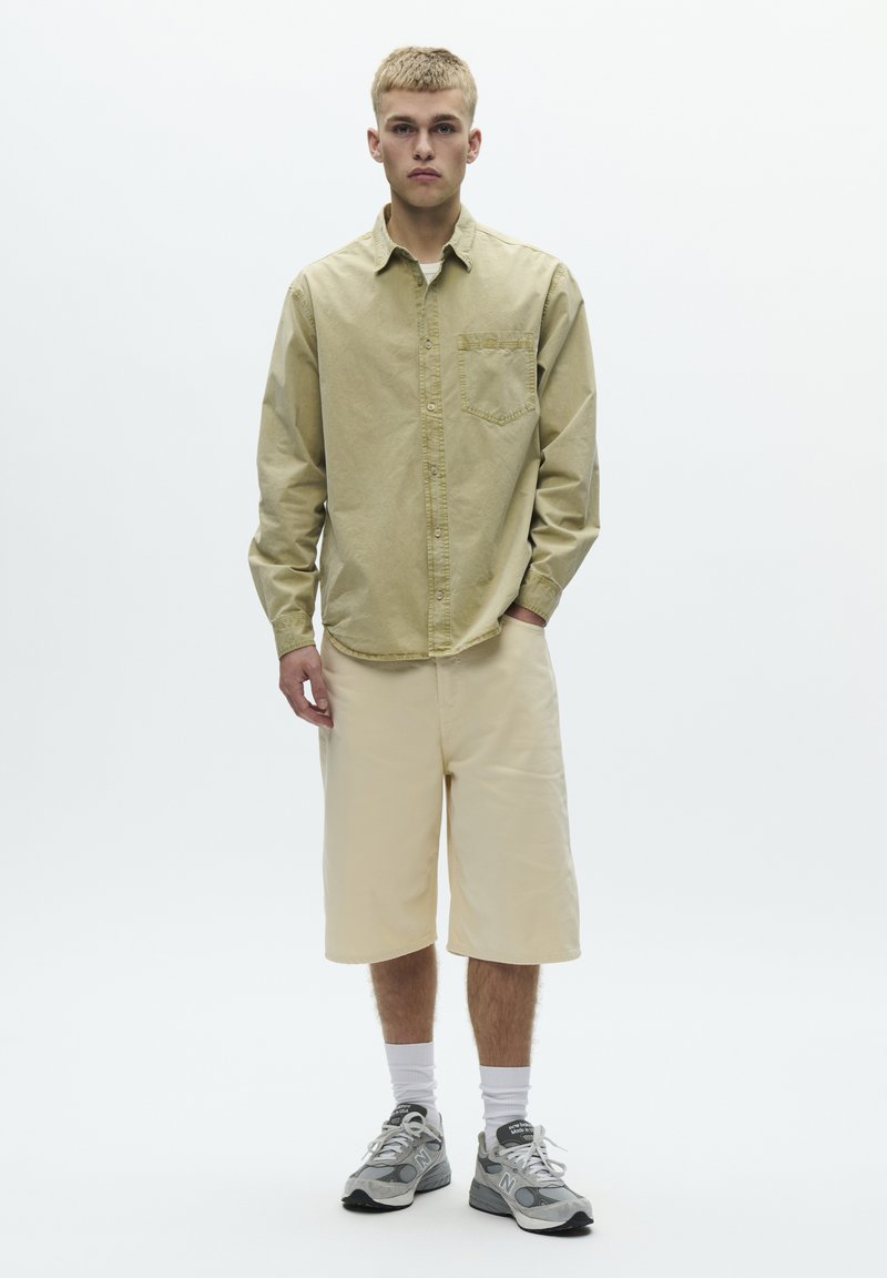Chemise à manches longues vert clair avec boutons, short beige jusqu'aux genoux, chaussures de sport grises et chaussettes blanches. Design propre et minimaliste.