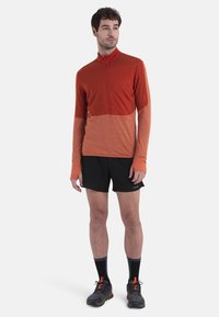 Maglia atletica a maniche lunghe in gradazioni di rosso, dotata di un colletto alto e mezza zip, abbinata a pantaloni corti neri e scarpe da corsa.