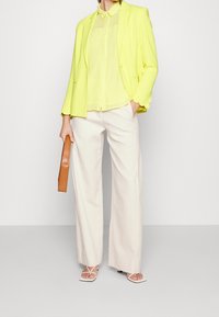 Blazer jaune fluo avec des manches en forme de vagues, superposé à un blouse verte transparente. Un pantalon large crème et une pochette marron complètent le look.