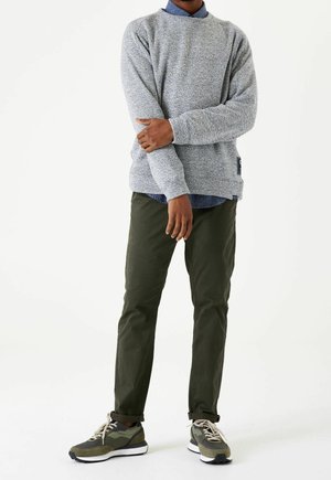 Mand iført grå tekstureret sweater, blå skjorte med krave, olivengrønne bukser og grønne og hvide sneakers, stående med armene over kors.