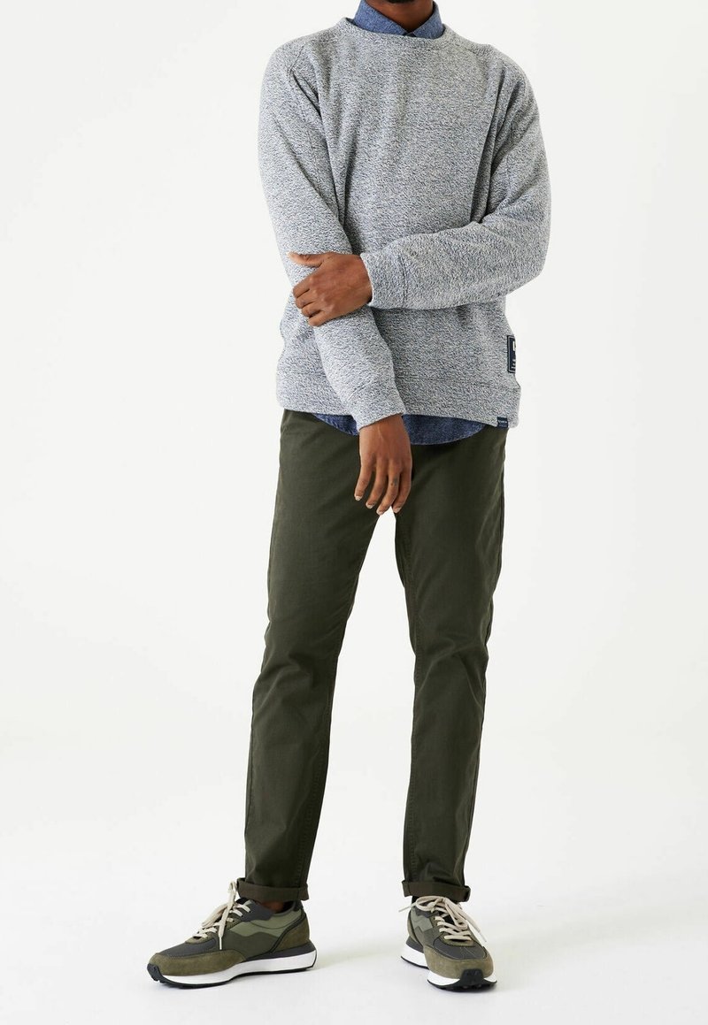 Homme portant un pull gris texturé, une chemise bleue à col, un pantalon vert olive et des baskets vertes et blanches, debout, les bras croisés.