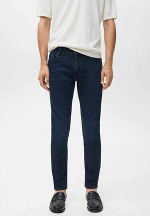 Una persona está de pie vistiendo una camisa de color claro y jeans oscuros, combinados con mocasines negros, frente a un fondo blanco liso.