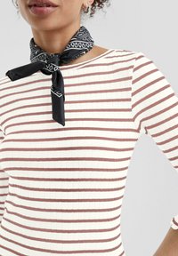 Geribbelde, aansluitende top met horizontale witte en bruine strepen. Accessoire: zwarte sjaal met witte patronen, om de nek geknoopt.