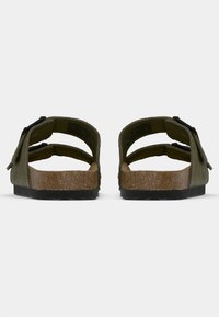 Olivnozeleni sandali s plitkim podplatom od plute, prilagodljivimi trakovi in črnimi dodatki. Oblikovanje vključuje teksturirano površino in ravno gumijasto podplato.