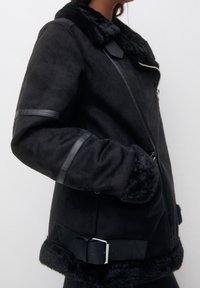 Veste noire en suédine synthétique avec un col et des poignets en fourrure. Comprend une fermeture éclair, des accents en cuir et une large ceinture à la taille.