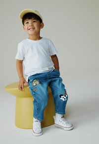 Siniset farkut, joissa on traktorin ja lehmän kirjailu, yhdistetty valkoiseen t-paitaan ja valkoisiin lenkkareihin. Lapsi, jolla on keltainen lippalakki, istuu keltaisella jakkaralla.