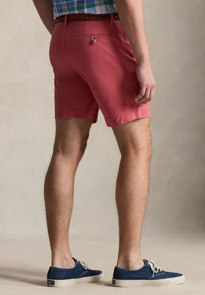 Κόκκινα βαμβακερά σορτς με πίσω τσέπη και λεπτομέρεια με κουμπί, σε συνδυασμό με ναυτικού χρώματος πάνινα αθλητικά παπούτσια. Το σκηνικό έχει ουδέτερο φόντο.