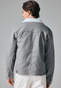 Grå denimjakke med hvid, fluffy krave. Har knaplukning ved ærmerne og lige snit, ensfarvet ryg med minimale syninger.