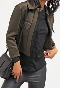 Giacca cropped verde oliva con colletto e due tasche frontali, abbinata a una camicia nera e una borsa nera elegante. L'outfit presenta un design moderno.