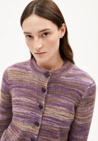 Jeune femme aux longs cheveux bruns portant un cardigan boutonné rayé violet et marron, regardant directement l'appareil photo sur un fond blanc.