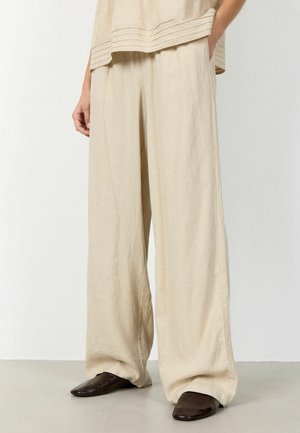 Beige Leinenhose mit weitem Bein, getragen mit einem passenden Oberteil und dunkelbraunen spitzen Schuhen, an einer stehenden Person vor einem schlichten Hintergrund.