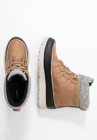 Braune Winterstiefel mit grauen Wollmanschetten, schwarzen Zehenkappen, gewickelten schwarz-weißen Sohlen und Schnürung auf weißem Hintergrund.