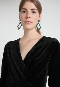 Rochie din catifea neagră, cu mâneci lungi; decolteu adânc în V; modelul poartă cercei verzi cu motive geometrice. Materialul texturat reflectă lumina subtil.