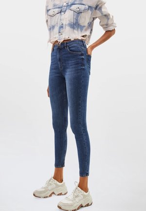 Kobieta w ciemnoniebieskich, wysoki stan, obcisłych jeansach, podwiniętej kurtce typu tie-dye oraz białych masywnych sneakersach, stojąca z jedną ręką w kieszeni.