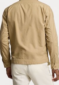 Muž v béžové dlouhé bundě a bílých kalhotách, zobrazený zezadu na pozadí s čistě bílou plochou.