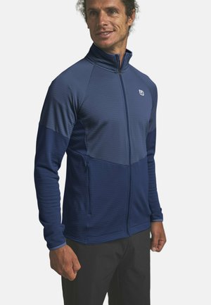 Mann trägt eine marineblaue Jacke mit durchgehendem Reißverschluss, hohem Kragen und Reißverschlusstaschen an den Seiten, steht vor einem einfarbigen Hintergrund.