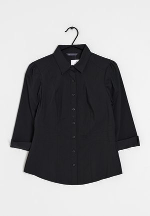 Sort langærmet bluse med knapper og krave samt let puffede skuldre, hængende på en sort bøjle mod en hvid baggrund.