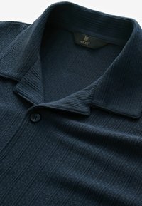 Polo de punto azul marino con una tela texturizada, que presenta un cuello acanalado y un único botón negro cerca de la tapeta. Etiqueta de la marca visible en el interior del cuello.