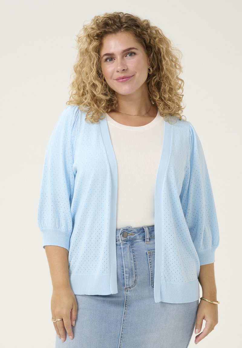 Cardigan en maille bleu clair à coupe ample, avec un motif texturé, des poignets côtelés et un décolleté en V, superposé sur une chemise blanche.