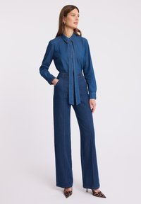 Kobieta ubrana w niebieską koszulę dżinsową z krawatem oraz spodnie z wysokim stanem i szerokimi nogawkami, pozująca z rękami w kieszeniach, w butach na obcasie w leopardski wzór.