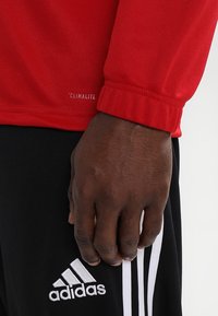 Rode textuurtrui met omslagmouwen, voorzien van een "CLIMALITE" label. Zwarte broek met drie witte strepen en een Adidas-logo op het been.