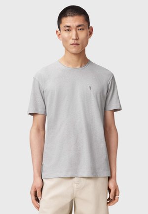 Jeune homme aux cheveux courts portant un t-shirt à manches courtes gris clair et un pantalon beige, debout devant un fond uni clair.