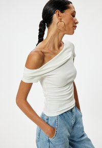 Witte geribbelde top met een off-the-shoulder ontwerp, voorzien van gedrapeerde details. Combi met lichtblauwe denim jeans, wat een casual uitstraling geeft.