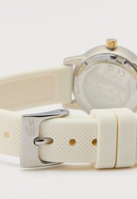 Άσπρο λαστιχένιο λουράκι ρολογιού με υφή και ασημί πόρπη χαραγμένη με λογότυπο κροκοδείλου, με θολωμένη ασημί κάσα ρολογιού στο φόντο.