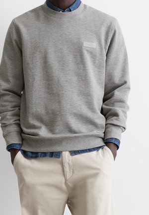 Homme portant un sweat-shirt gris clair sur une chemise bleue et un pantalon beige, les mains dans les poches, sur fond blanc.