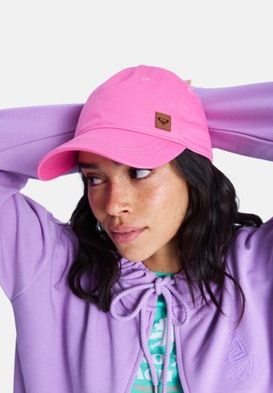 Woman wearing a pink cap and lavender hoodie with drawstrings, looking to her left with hands behind her head against a white background.