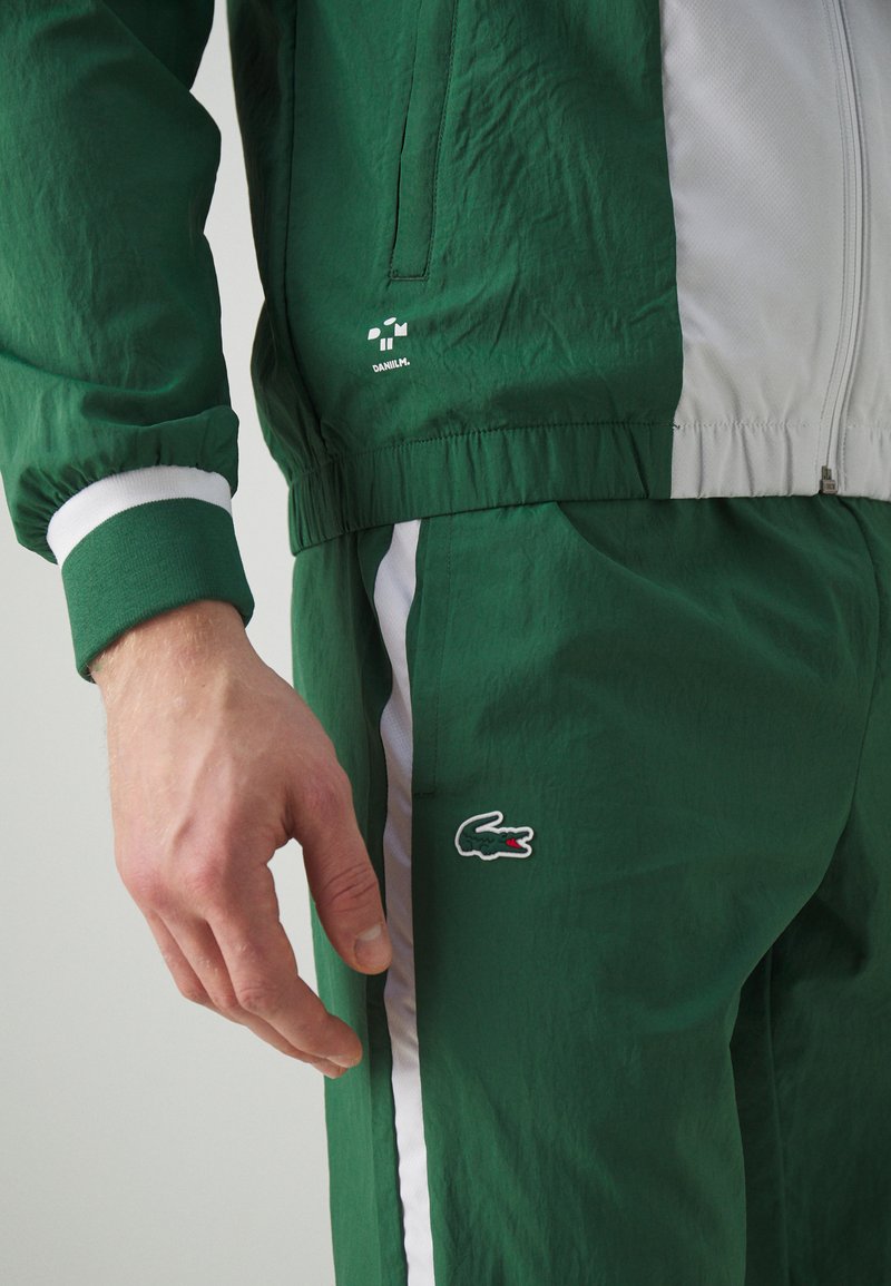Zielona kurtka sportowa z białymi akcentami, elastycznymi mankietami i lekką teksturą; w połączeniu z zielonymi spodniami z bocznymi paskami i detalem logo.