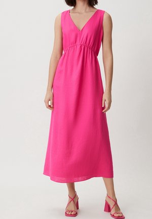 Vrouw die een mouwloze midi-jurk met V-hals in felroze draagt en bijpassende sandalen met blokhakken en bandjes, staand tegen een effen achtergrond.
