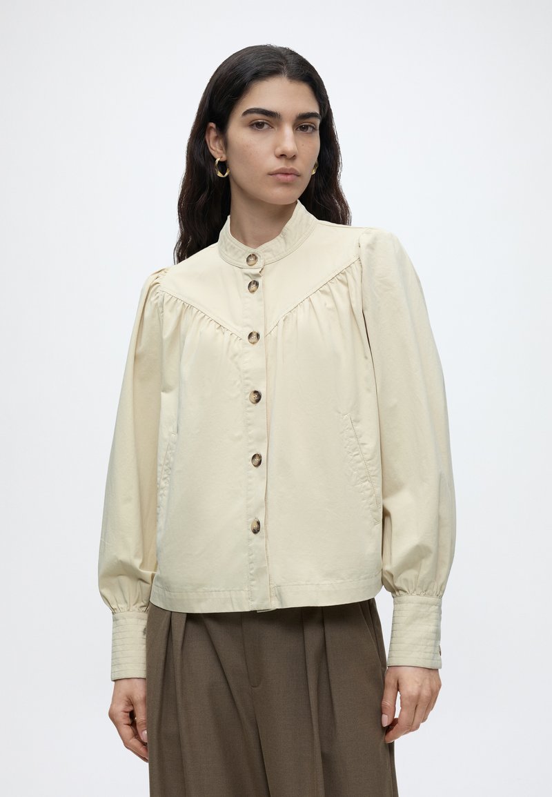 Femme aux longs cheveux foncés portant des boucles d'oreilles créoles dorées, une blouse beige boutonnée avec des manches bouffantes et un pantalon large marron sur fond blanc.