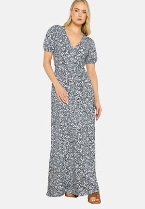 Donna che indossa un abito lungo a fantasia floreale bianco e nero con scollo a V, maniche corte a sbuffo e cintura annodata in vita, abbinato a sandali marroni.